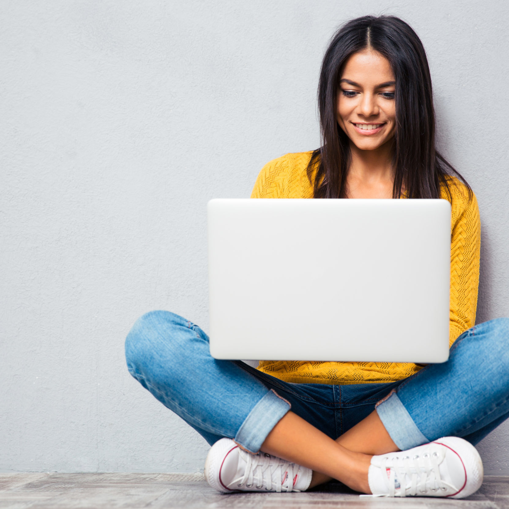 Happy young woman sitting on the floor with corssed legs and using laptop on gray background